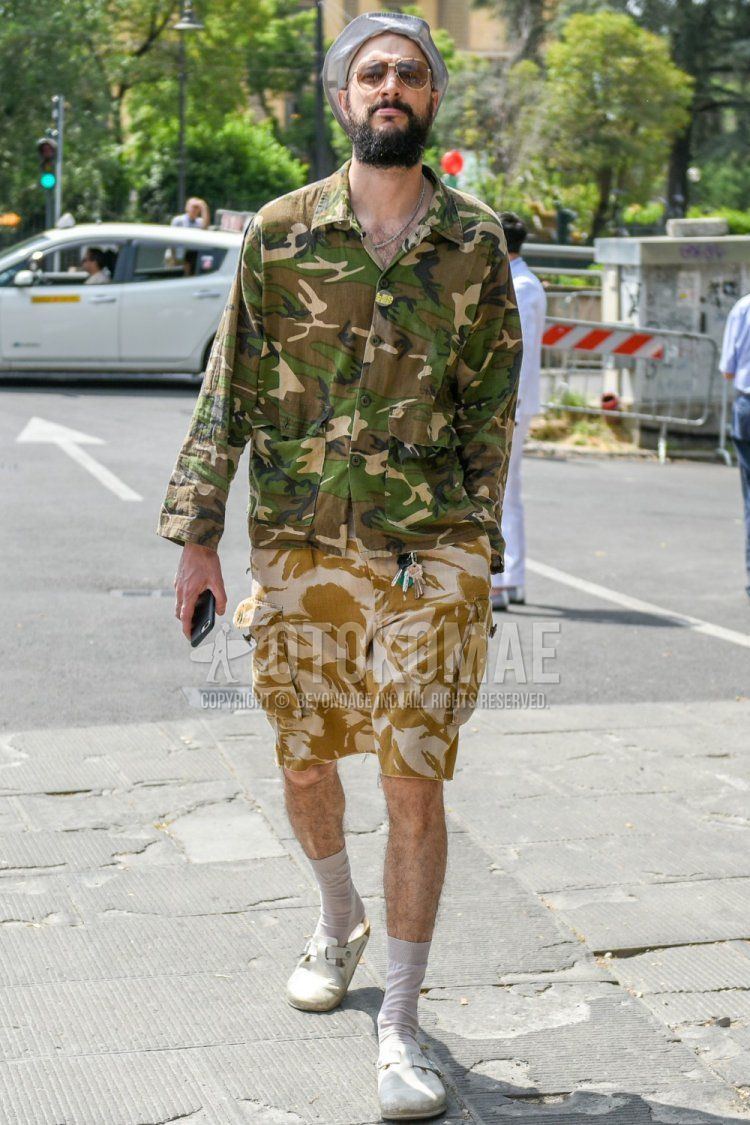 Spring and summer men's coordinate and outfit with bucket solid gray hat, teardrop solid gold-brown sunglasses, olive green camouflage shirt, beige bottoms cargo pants, bottom shorts, solid white socks, and white leather sandals.