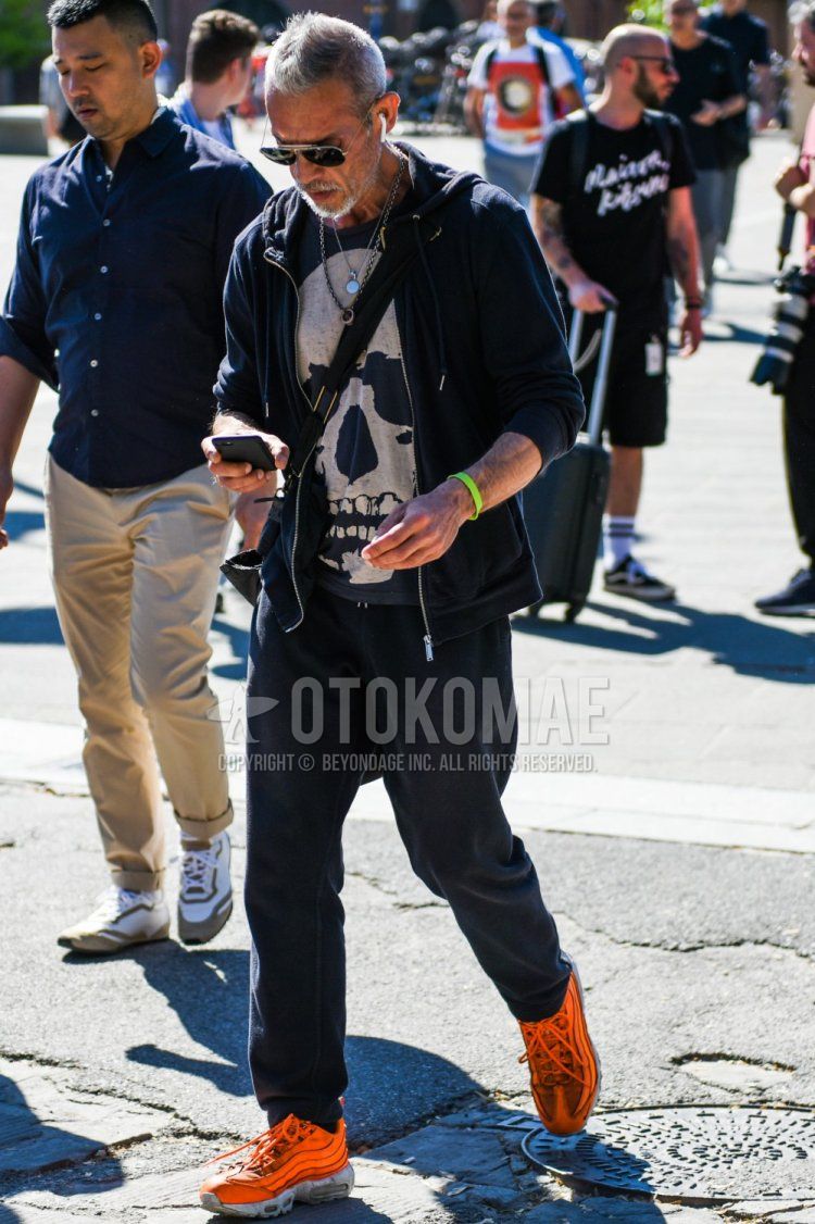 Spring and fall men's coordinate and outfit with teardrop black and silver plain sunglasses, plain black hoodie, black graphic t-shirt, plain black sweatpants, plain jogger pants/ribbed pants, and Nike Air Max 95 orange low-cut sneakers.