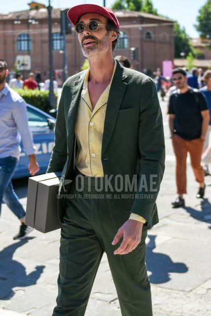 Men's spring/summer coordinate and outfit with red solid color baseball cap, round gold/black solid color sunglasses, open collar yellow solid color shirt, and olive green solid color suit.