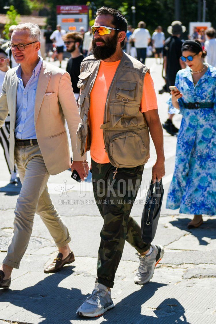 Men's spring/summer outfit with plain white/multicolor sunglasses, solid beige gilet, solid orange t-shirt, green camouflage cotton pants, green low-cut sneakers, and solid black briefcase/handbag.