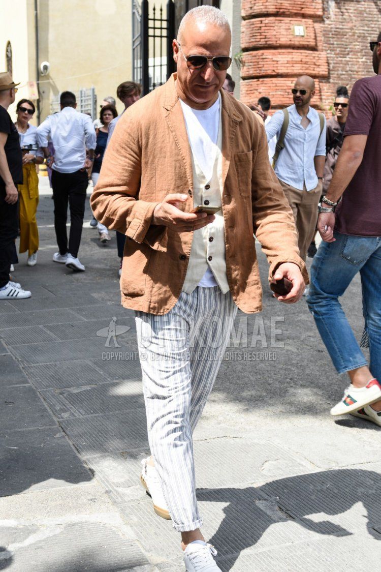Men's spring, summer, and fall coordinate and outfit with plain silver sunglasses, plain brown tailored jacket, plain white T-shirt, plain white gilet, white/gray striped cotton pants, and white low-cut sneakers.