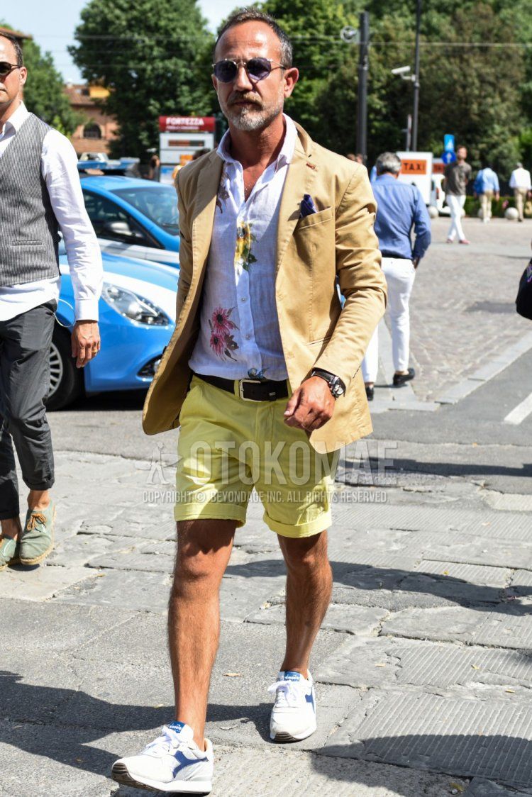 Men's spring/summer coordinate and outfit with plain silver sunglasses, plain beige tailored jacket, linen white botanical shirt, plain black leather belt, plain yellow shorts, and white low-cut Diadora sneakers.