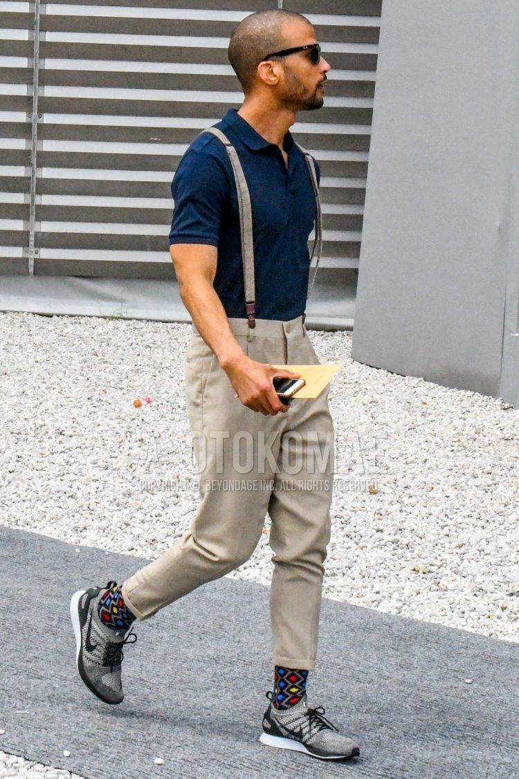 Men's summer coordinate and outfit with solid color sunglasses, solid color navy polo shirt, solid color beige suspenders, solid color beige cropped pants, multi-colored socks socks, and Nike gray low-cut sneakers.