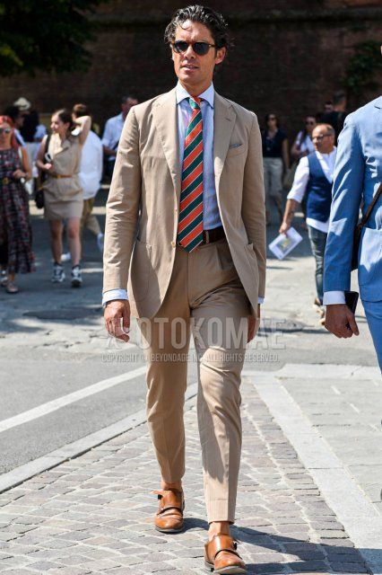 Men's spring, summer, and fall coordination and outfit with plain brown/black sunglasses, plain light blue shirt, plain brown leather belt, brown monk shoe leather shoes, plain beige suit, and orange/green regimental tie.