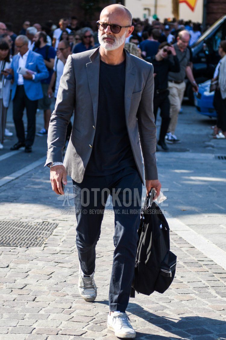 Men's spring, summer, and fall coordinate and outfit with brown tortoiseshell sunglasses from Boston, plain gray tailored jacket, plain black t-shirt, plain black slacks, Philip model white low-cut sneakers, and plain black backpack.