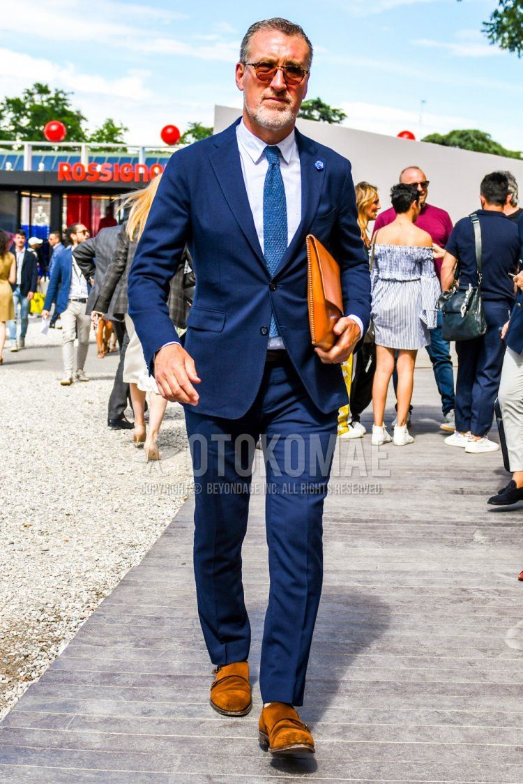 Men's spring/summer/fall outfit with solid color sunglasses, solid color white shirt, brown monk shoes leather shoes, brown wingtip leather shoes, solid color brown clutch bag/second bag/drawstring, solid color navy suit, solid color blue knit tie.