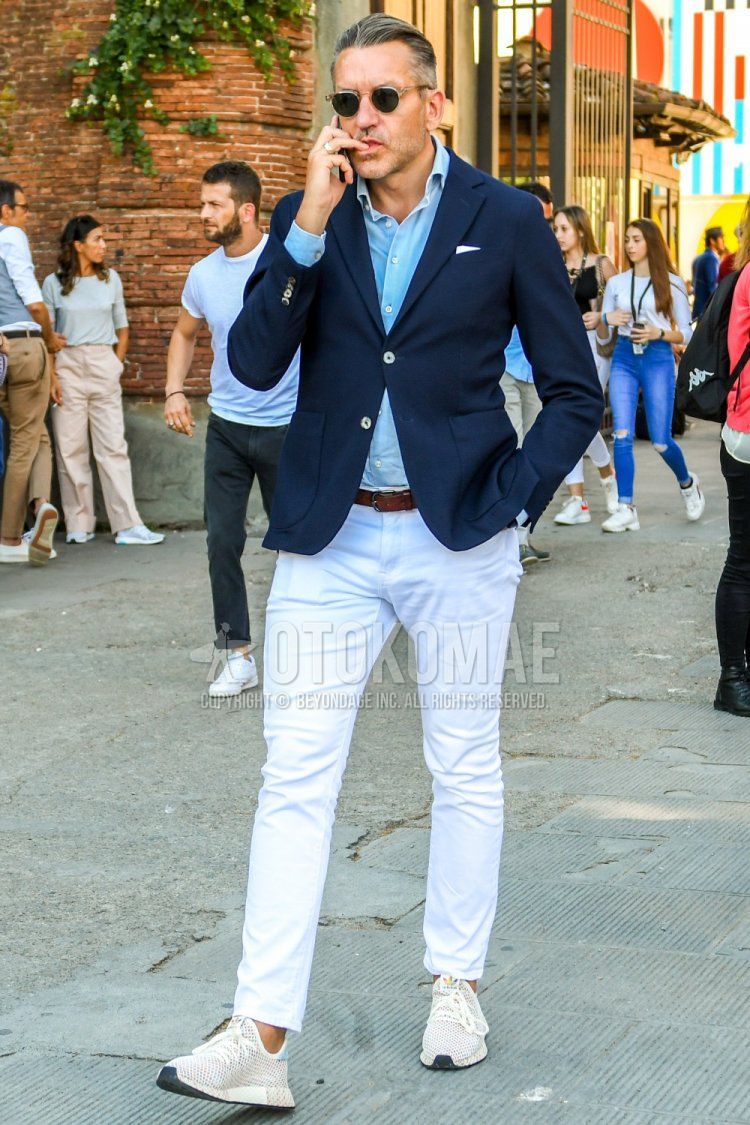 Men's spring, summer, and fall coordinate and outfit with solid color sunglasses, solid color navy tailored jacket, solid color light blue shirt, solid color brown leather belt, solid color white ankle pants, and white low-cut sneakers.