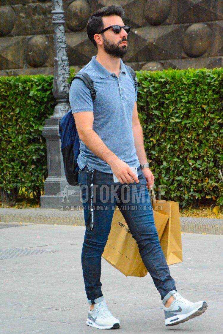 Men's spring/summer outfit with Wellington plain black sunglasses, plain gray polo shirt, plain navy denim/jeans, Nike gray low-cut sneakers, and plain gray/black backpack.