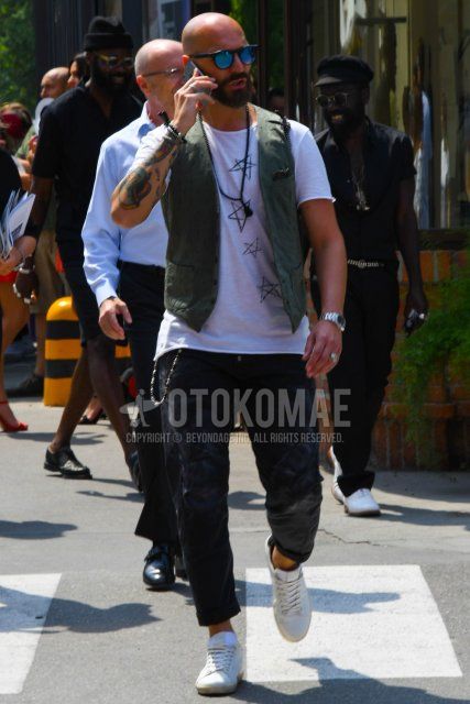 A summer men's coordinate and outfit with plain sunglasses, a plain olive green gilet, a white graphic t-shirt, plain black cotton pants, and white low-cut sneakers.