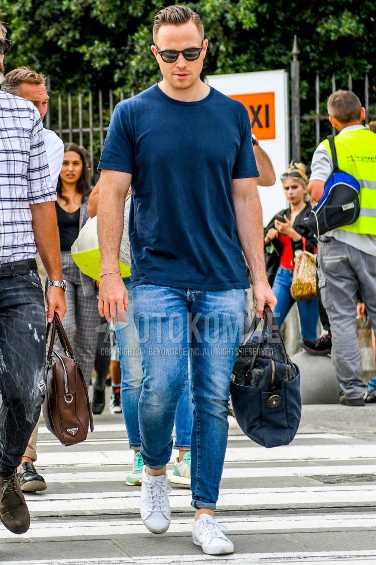 Summer men's coordinate and outfit with brown tortoiseshell sunglasses, plain navy t-shirt, plain blue denim/jeans, white low-cut sneakers, and plain navy briefcase/handbag.