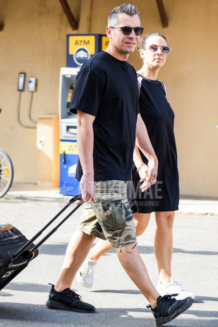 Summer men's coordinate and outfit with plain black sunglasses, plain black t-shirt, olive green camouflage cargo pants, camouflage shorts, and black low-cut sneakers.