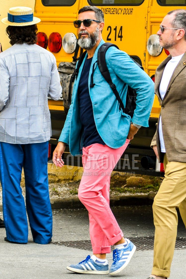 Men's spring/summer/fall outfit with plain navy sunglasses, plain light blue tailored jacket, plain navy t-shirt, plain pink cotton pants, Adidas blue low-cut sneakers, and plain black backpack.