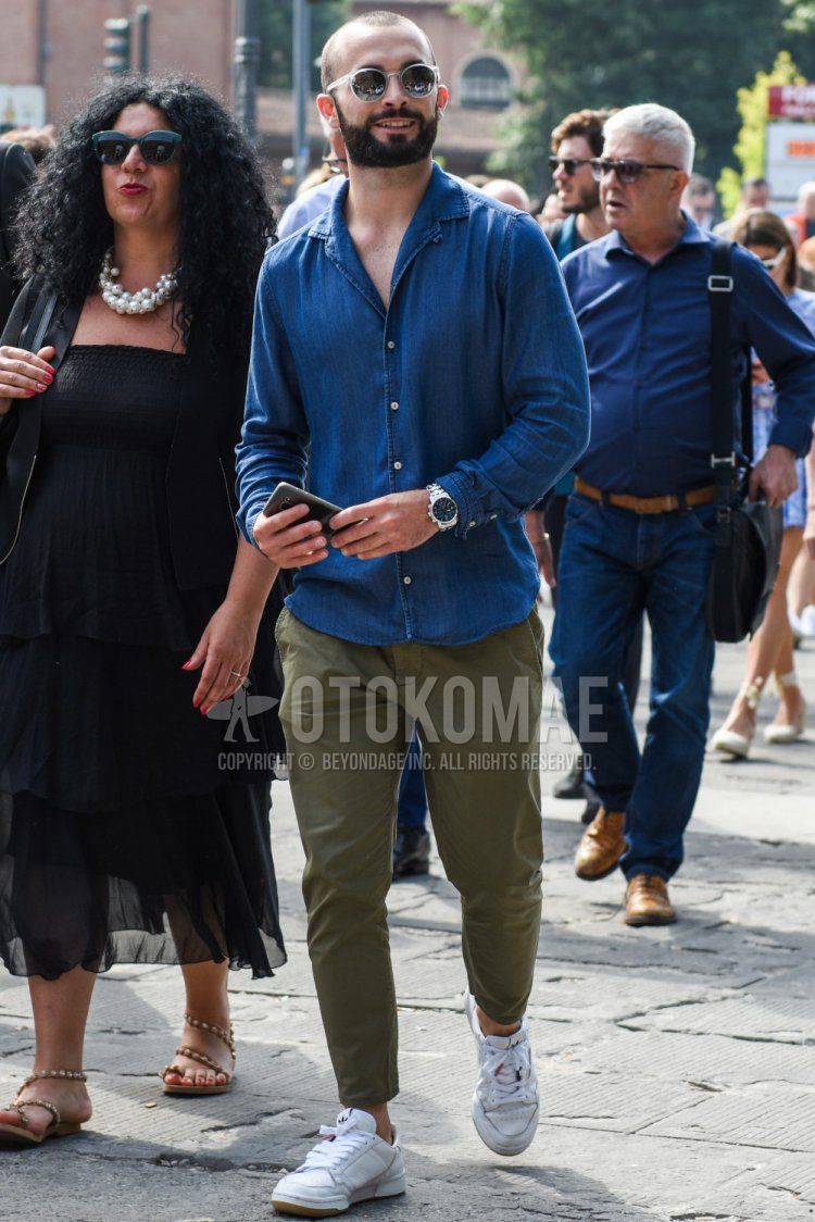 Men's spring/summer coordinate and outfit with clear/white solid color sunglasses, blue solid color denim/chambray shirt, olive green solid color chinos and white low-cut Adidas sneakers.