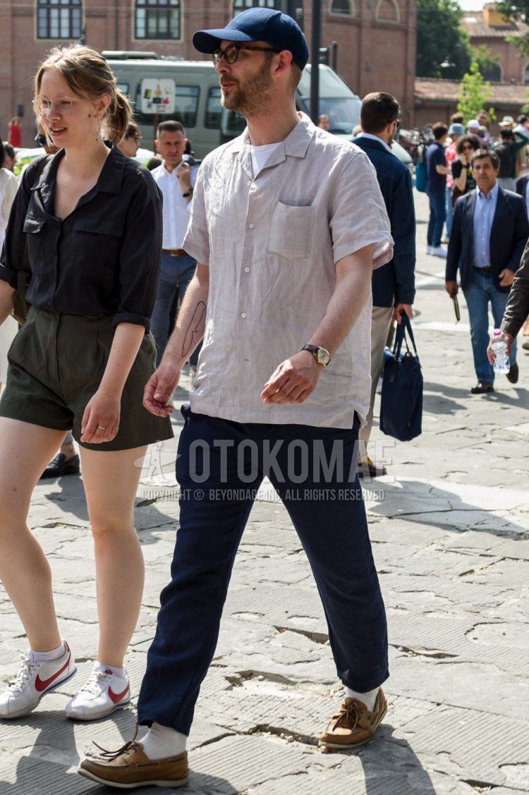 Summer men's coordinate and outfit with plain navy baseball cap, brown tortoiseshell glasses, plain beige shirt, plain white t-shirt, plain navy ankle pants, plain white socks, and beige moccasins/deck shoes leather shoes.