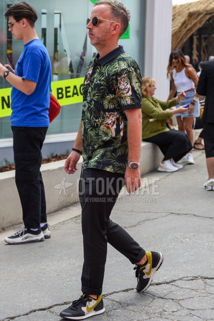 Men's spring and summer coordinate and outfit with round gold solid color sunglasses, black/green botanical polo shirt, dark gray solid color slacks, and Nike black/beige low-cut sneakers.