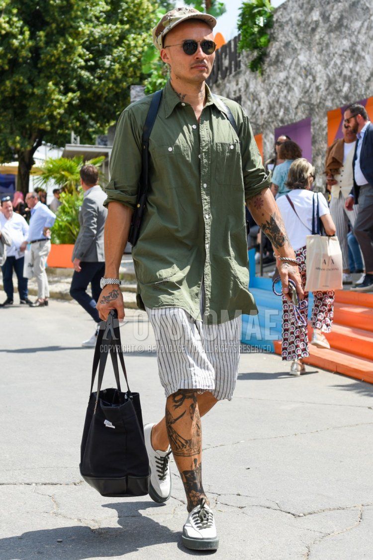 Summer men's coordinate and outfit with beige baseball cap, round solid black sunglasses, olive green solid shirt, white striped shorts, white low-cut sneakers, and solid black tote bag.