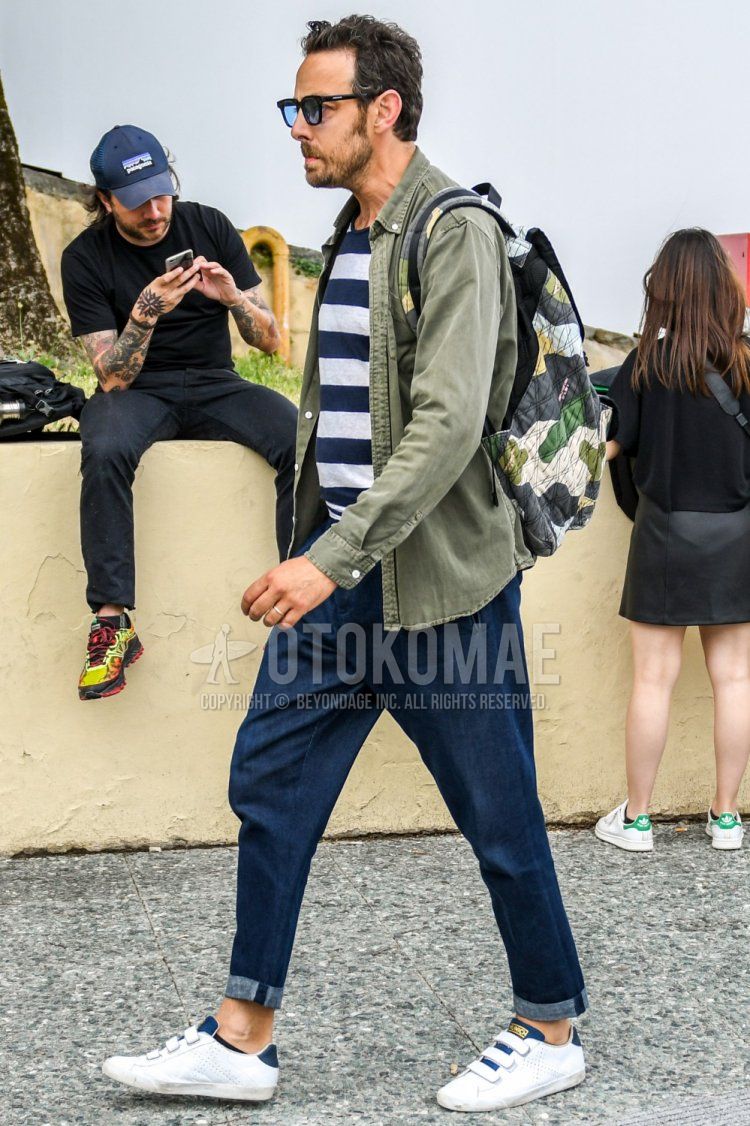 Men's spring/summer/fall coordinate/outfit with plain sunglasses, plain olive green shirt, navy/white striped t-shirt, plain navy denim/jeans, white low-cut sneakers, and multi-colored camouflage backpack.