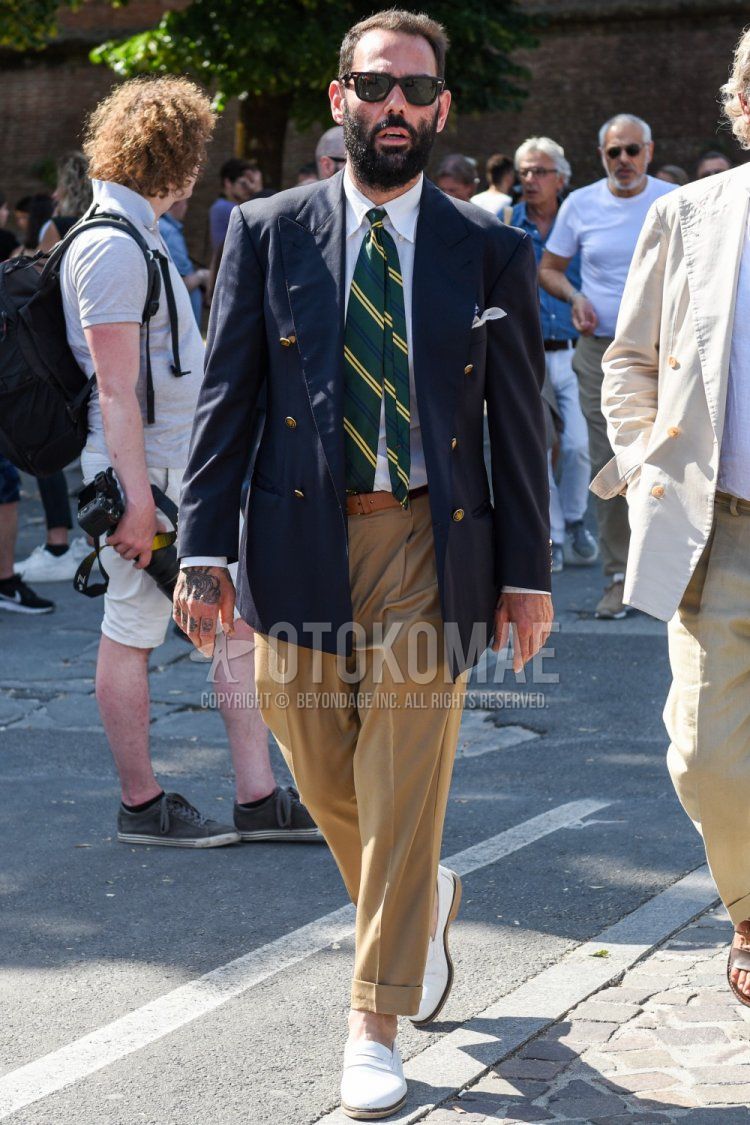 Men's spring and fall outfit with brown tortoiseshell sunglasses, plain navy tailored jacket, plain white shirt, plain brown leather belt, plain beige chinos, white coin loafer leather shoes, and green regimental tie.