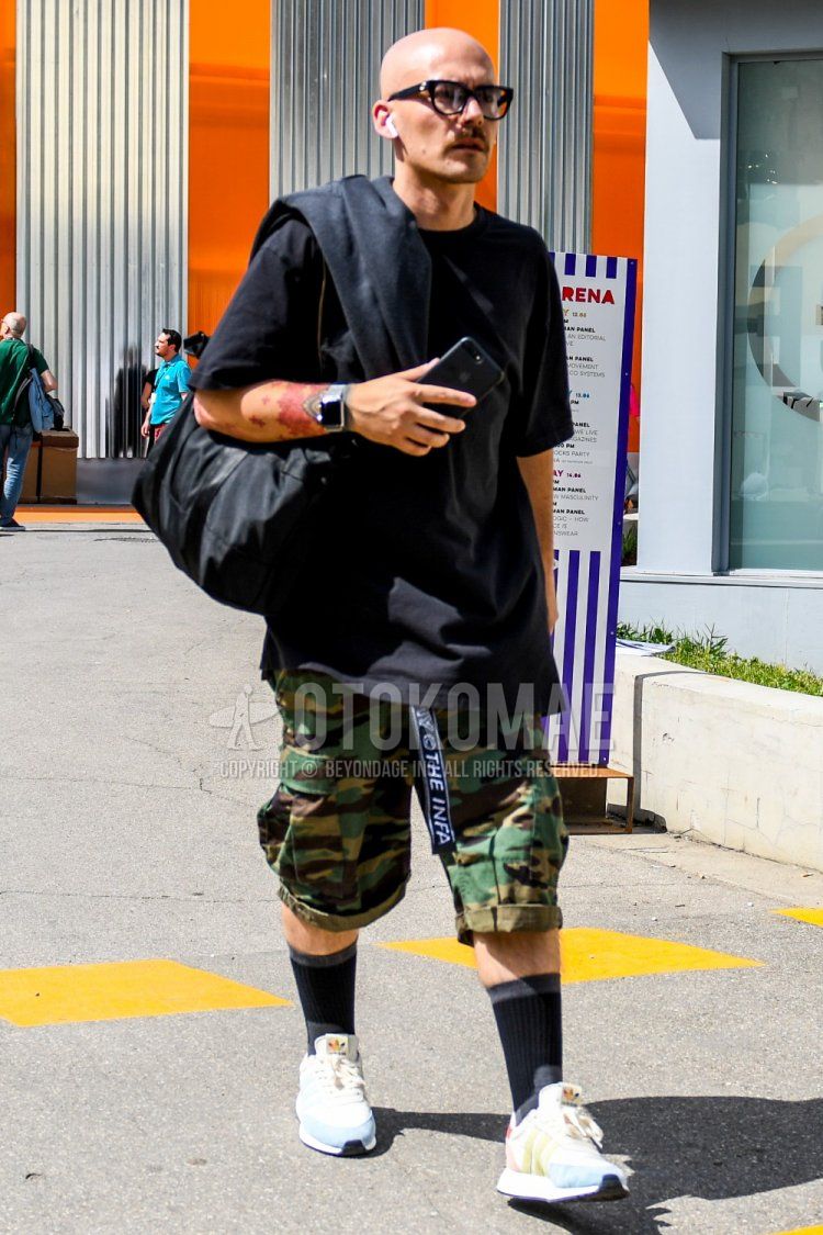 Men's summer coordinate and outfit with plain glasses, plain black t-shirt, white graphic tape belt, multi-colored camouflage cargo pants, multi-colored camouflage shorts, black/dark gray socks, and white low-cut Adidas sneakers.