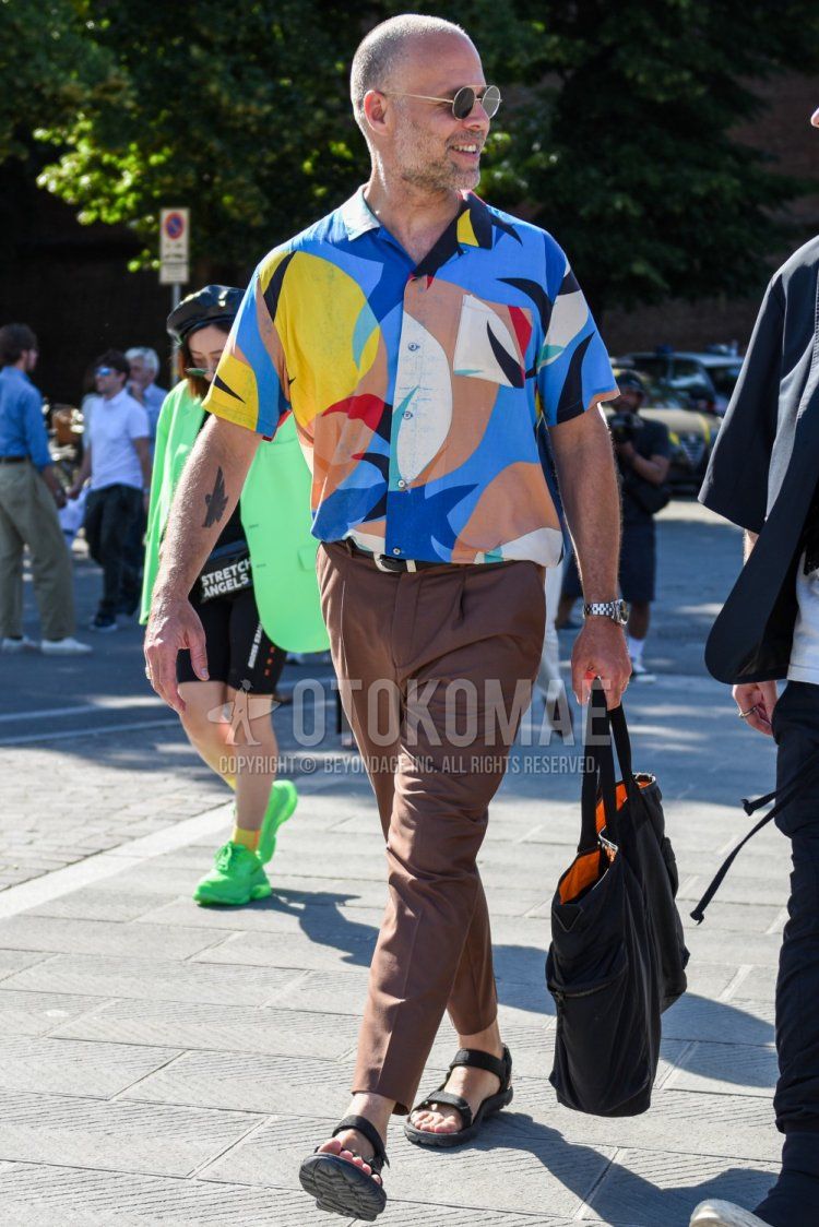 Men's summer coordinate and outfit with plain gold sunglasses, multi-colored top/inner shirt, plain brown slacks, black sport sandals, and plain black tote bag.