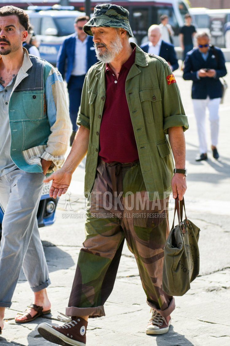Men's spring/summer/fall outfit with green camouflage hat, olive green solid shirt jacket, red solid polo shirt, beige/olive green solid cargo pants, brown high-cut sneakers, and olive green solid Boston bag.