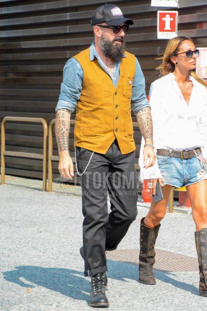 Men's spring/summer/autumn coordination and outfit with black one-pointed baseball cap, plain black Tom Ford sunglasses, plain yellow gilet, plain light blue denim/chambray shirt, plain black chinos, and black boots.