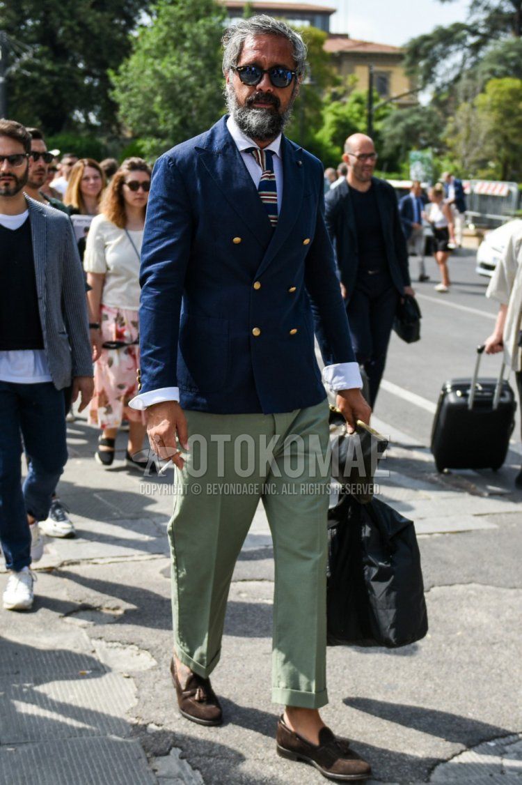 Men's spring/fall outfit with beige/black tortoiseshell sunglasses, plain navy tailored jacket, plain white shirt, plain green chinos, suede brown tassel loafer leather shoes, plain black briefcase/handbag, navy striped knit tie.