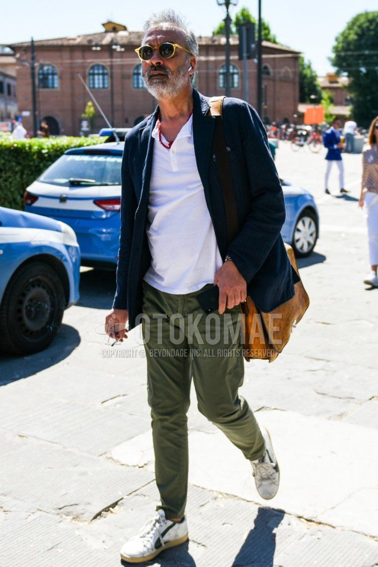 Men's spring/summer/fall outfit with clear/beige solid sunglasses, navy solid tailored jacket, white solid t-shirt with henley neck, olive green solid chinos, white low-cut sneakers, and beige solid shoulder bag.