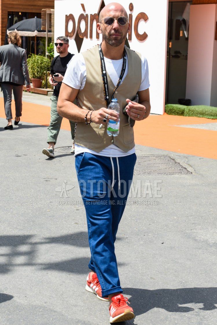Men's summer coordinate and outfit with plain silver sunglasses, plain beige gilet, plain white t-shirt, plain blue easy pants, and Adidas red low-cut sneakers.