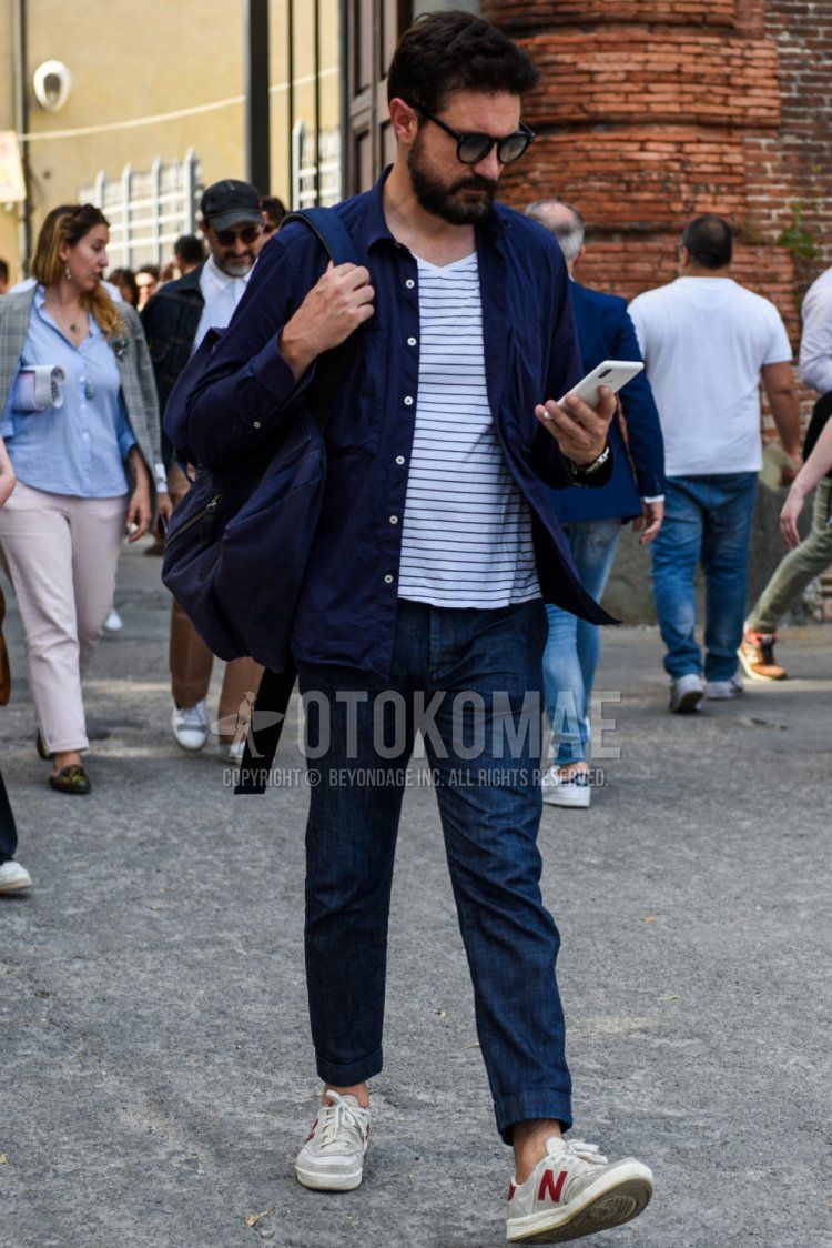 Men's spring, summer, and fall coordinate and outfit with plain black sunglasses, plain navy shirt, white and black striped t-shirt, plain navy damaged jeans, and New Balance gray and white low-cut sneakers.