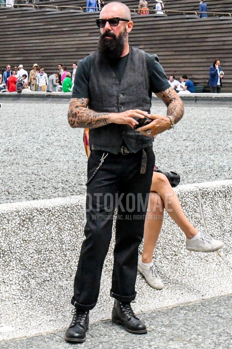Men's spring/fall outfit with plain black Tom Ford sunglasses, plain black t-shirt, plain gray gilet, plain black leather belt, plain black denim/jeans, and black work boots.