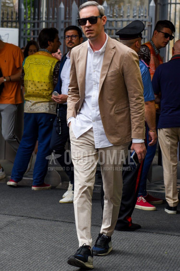 Men's spring/summer coordinate and outfit with plain black sunglasses, plain beige tailored jacket, plain white shirt, plain beige chinos, and black/blue low-cut sneakers.