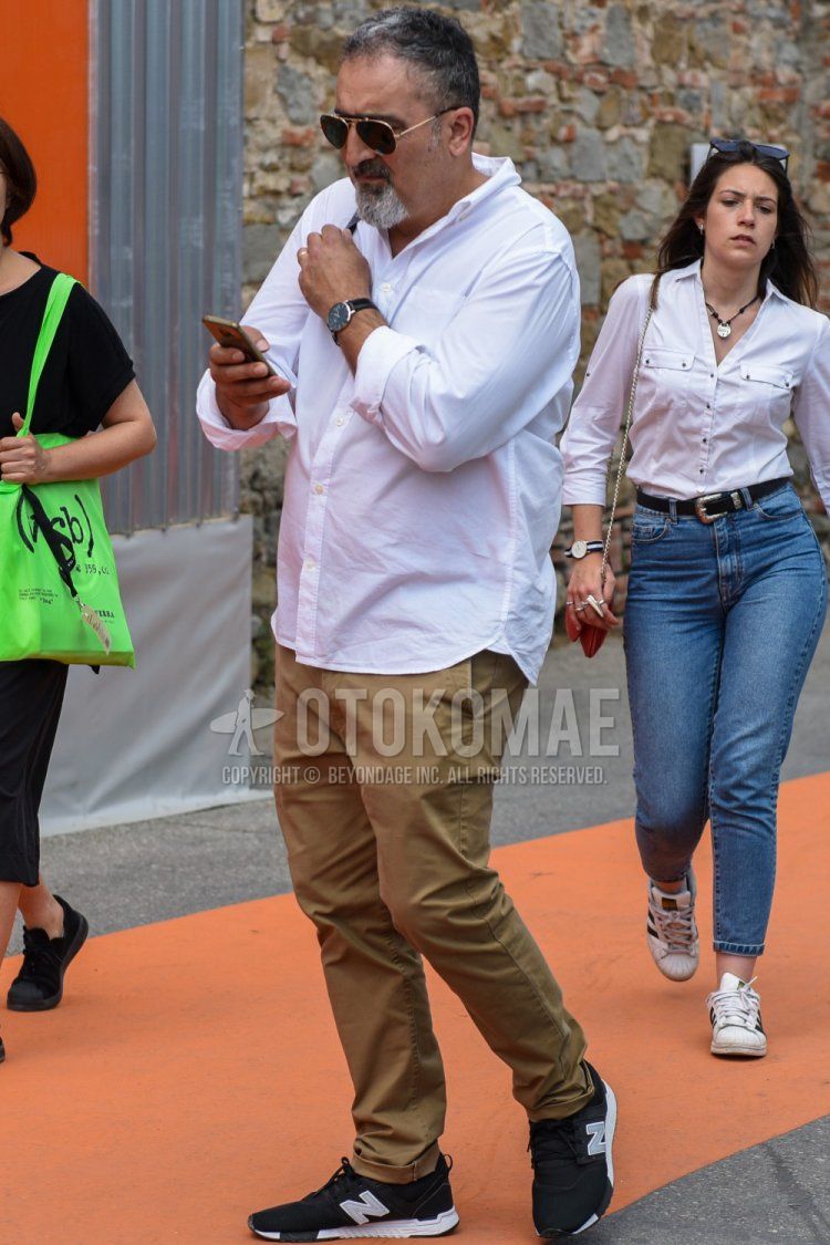 Men's spring, summer, and fall coordinate and outfit with gold and black plain sunglasses, white plain shirt, beige plain cotton pants, and black low-cut New Balance sneakers.