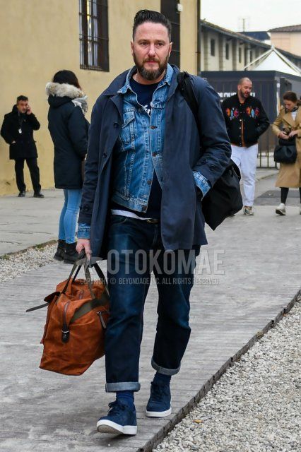 Men's fall/winter outfit with solid blue denim jacket, solid navy stainless coat, solid navy t-shirt, solid blue denim/jeans, solid gray socks, blue low-cut sneakers, and solid brown Boston bag.