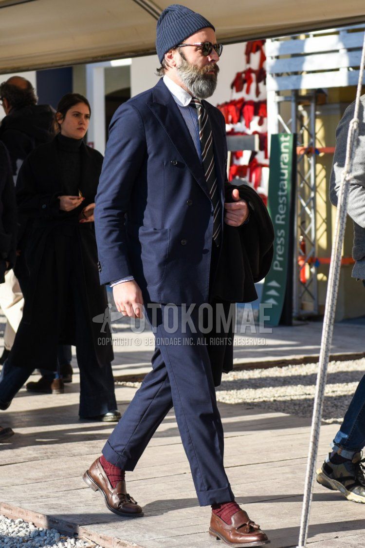 Men's spring and fall outfit with plain navy knit cap, brown tortoiseshell sunglasses, light blue striped shirt, solid red socks, brown tassel loafer leather shoes, plain navy suit, brown and black regimental tie.