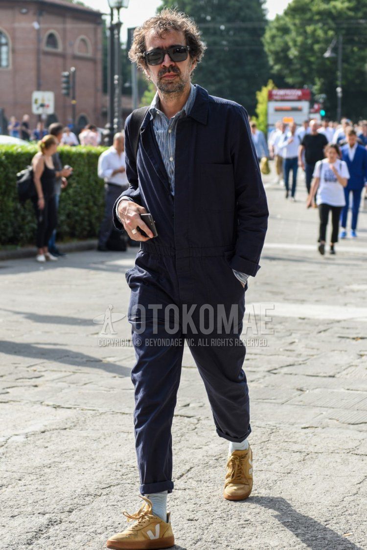 Men's spring and fall coordinate and outfit with square plain black sunglasses, plain navy jumpsuit, blue striped shirt, plain white socks, and beige low-cut sneakers.