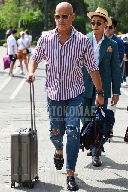 Men's spring/summer coordinate and outfit with teardrop gold/black solid sunglasses, white/red striped shirt, solid blue damaged jeans, black bit loafer leather shoes, solid blue/black backpack, solid gray suitcase.
