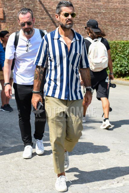 Summer men's coordinate and outfit with teardrop black and gold solid sunglasses, white and blue striped shirt, solid beige ankle pants, and white low-cut sneakers.