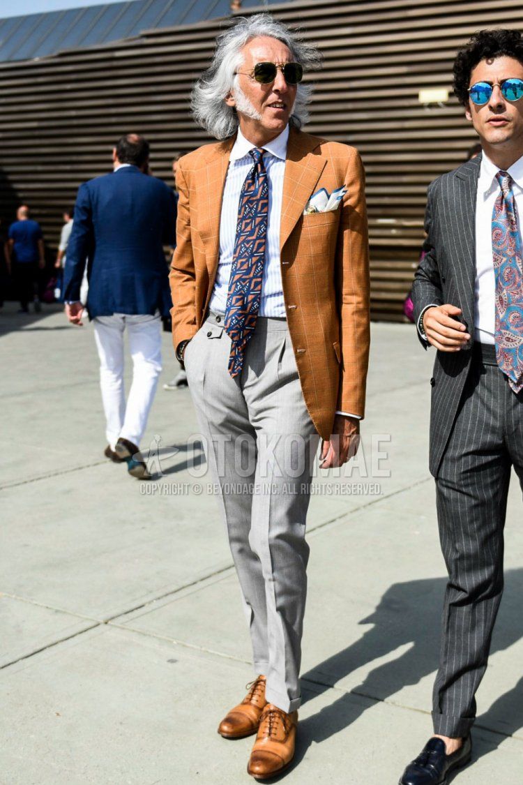 Men's spring and fall outfit with plain sunglasses, brown checked tailored jacket, white striped shirt, plain gray pleated pants, plain beltless pants, beige straight tip leather shoes, and orange/navy tie.