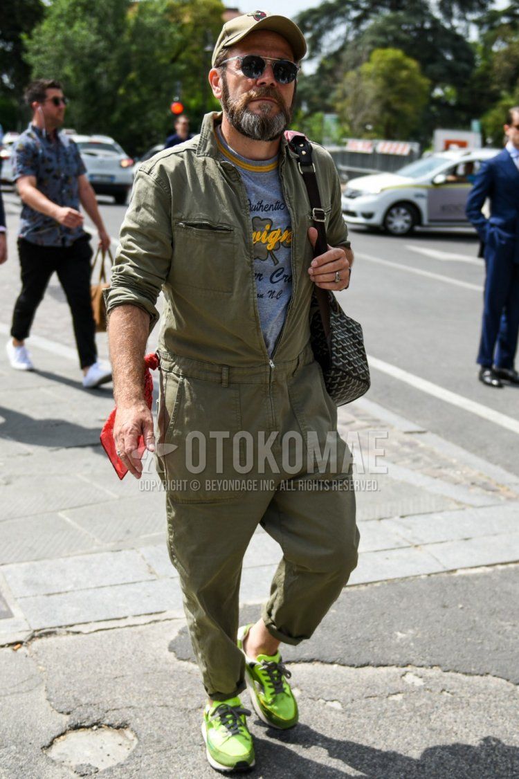 Men's spring, summer, and fall outfit with beige lettered baseball cap, solid black and gray sunglasses, solid olive green jumpsuit, gray graphic t-shirt, green low-cut sneakers, and Goyard black and gray bag shoulder bag.