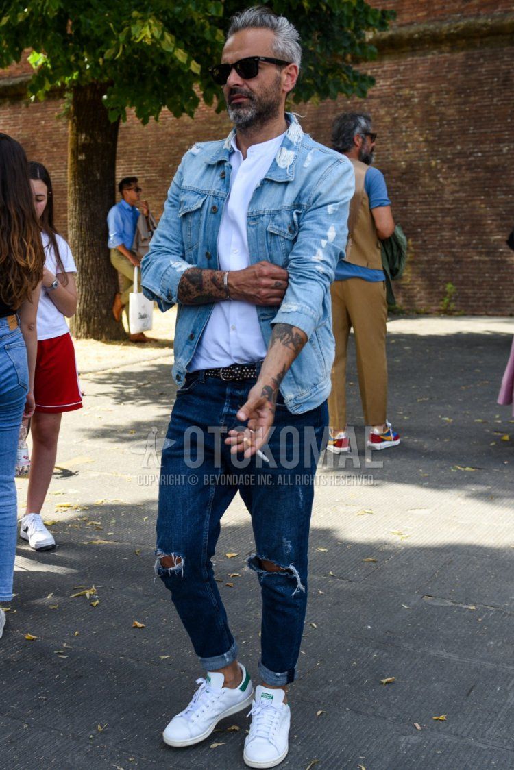 Men's spring, summer, and fall outfit with square solid black sunglasses, solid light blue denim jacket, solid white shirt, solid black leather belt, solid blue damaged jeans, and Adidas Stan Smith white low-cut sneakers.