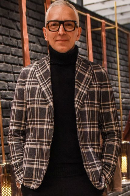Men's spring and fall outfit with Tom Ford brown tortoiseshell glasses, gray checked tailored jacket, solid black turtleneck knit, solid gray slacks, solid gray ankle pants, and white low-cut sneakers.