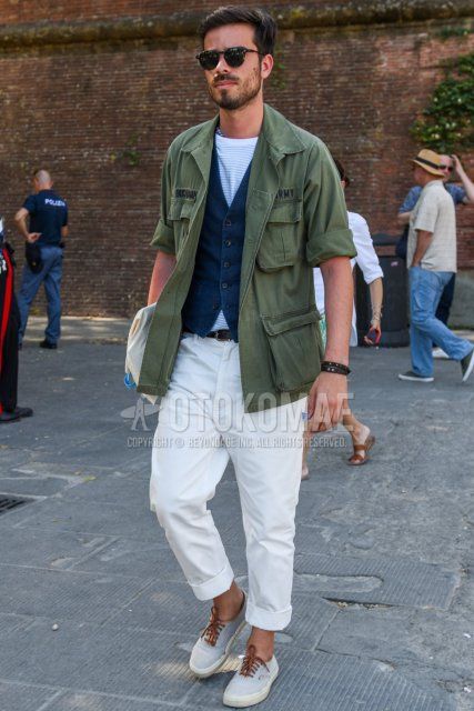 Men's spring and fall outfit with plain black sunglasses, plain olive green safari jacket, plain navy gilet, white striped t-shirt, plain white cotton pants, and white low-cut sneakers.