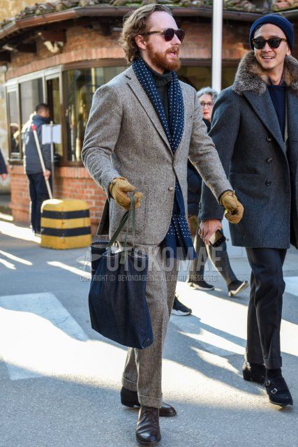 Men's spring/fall outfit with brown tortoiseshell sunglasses, navy-dot scarf/stall, plain gray turtleneck knit, brown chukka boots, plain navy briefcase/handbag, and plain gray suit.