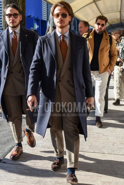 Men's fall/winter outfit with plain silver sunglasses, gray checked chester coat, light blue striped shirt, navy socks socks, suede brown tassel loafers leather shoes, plain gray suit, and multi-colored tie.