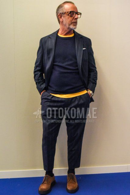 Men's spring and fall coordinate and outfit with brown tortoiseshell glasses, solid navy sweater, solid yellow t-shirt, solid black socks, suede brown plain toe leather shoes, and dark gray striped suit.