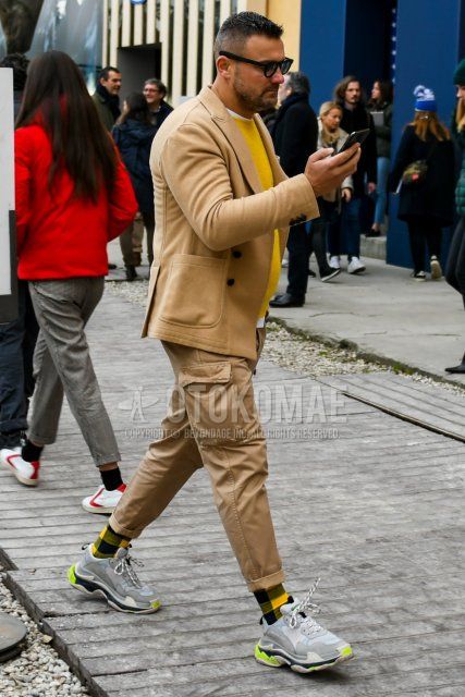 Men's spring and fall outfit with plain black sunglasses, plain beige tailored jacket, plain yellow sweater, plain white t-shirt, plain beige cargo pants, yellow/black checked socks, Balenciaga Triple S gray low-cut sneakers.