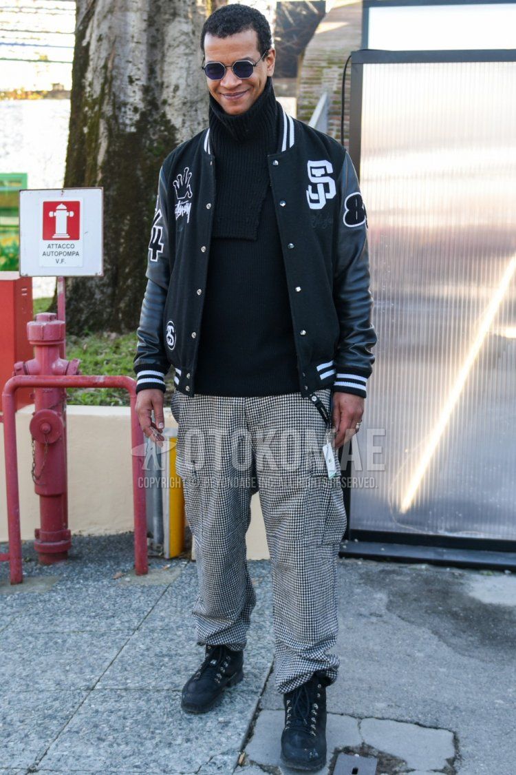 Men's fall/winter outfit and outfit with plain silver sunglasses, Stussy black lettered stadium jacket, plain black turtleneck knit, white/black checked slacks, white/black checked cargo pants, and black boots.