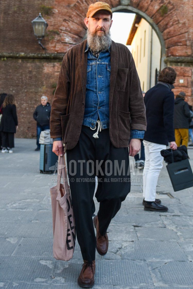 Men's fall/winter outfit and outfit with solid beige cap, solid brown outerwear, solid blue denim jacket, solid black easy pants, brown boots, and pink graphic tote bag.