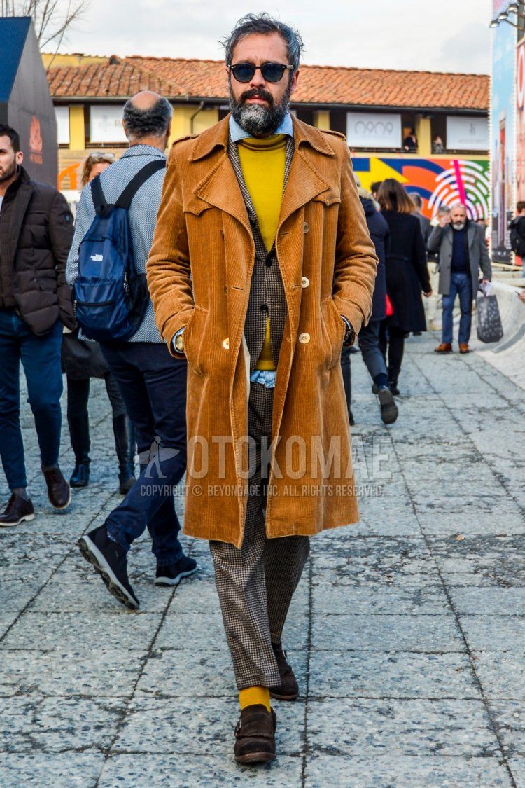 Men's fall/winter outfit/clothing with solid color sunglasses, solid color brown trench coat, solid color yellow sweater, solid color light blue denim/chambray shirt, brown monk shoes leather shoes, suede shoes leather shoes, brown checked suit.